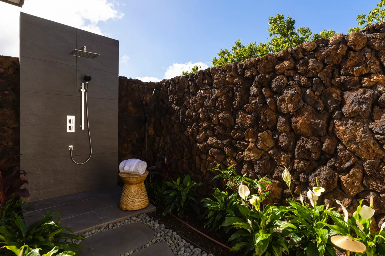 Una ducha exterior junto a un muro de piedra oscura y plantas frondosas.