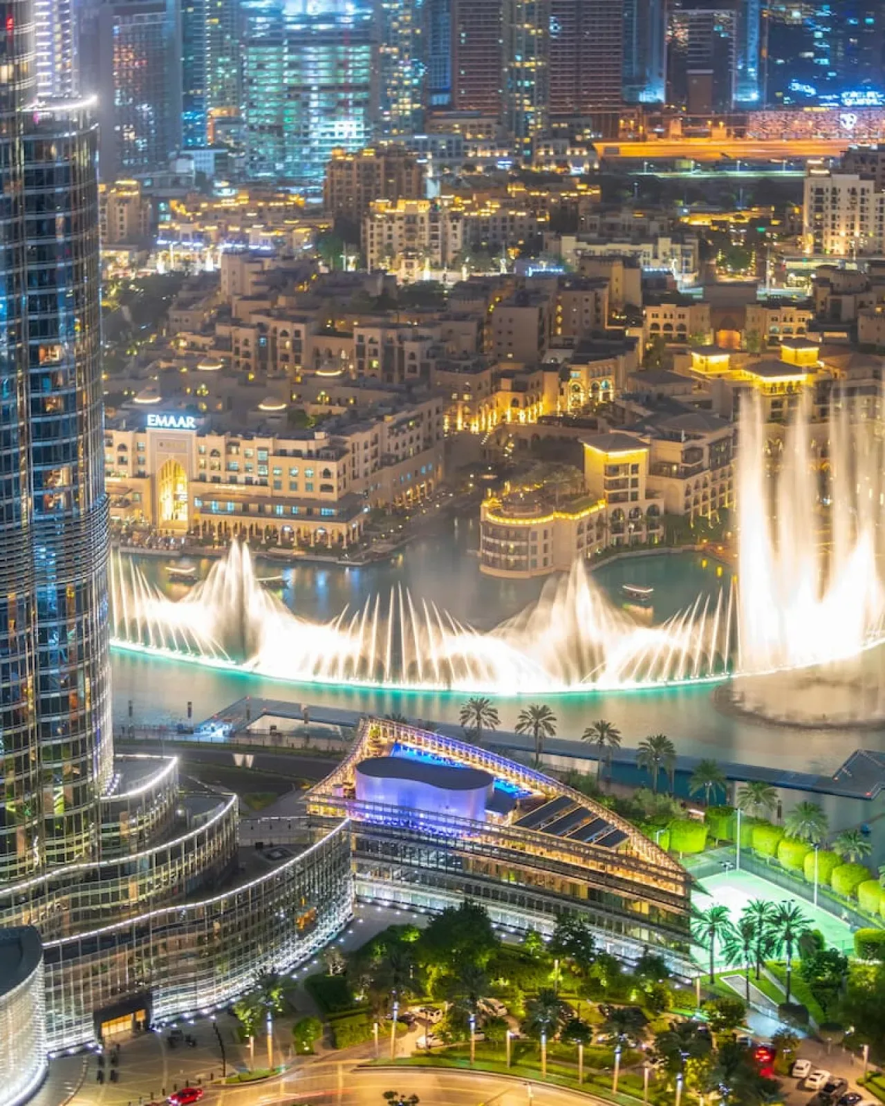 Un horizonte urbano nocturno domina fuentes iluminadas y un paseo junto al agua.
