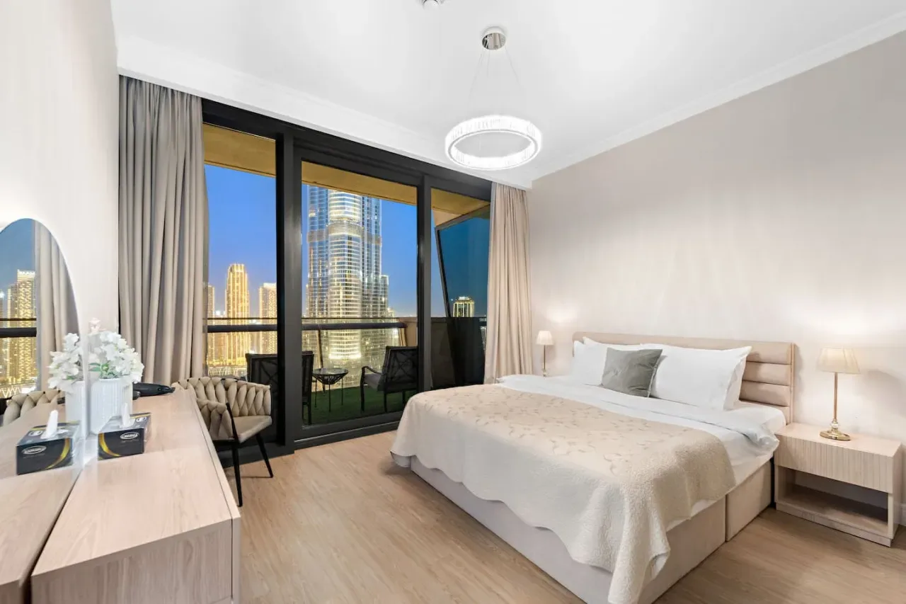 Un dormitorio luminoso con una cama tendida y ventanales que dan a edificios de la ciudad.
