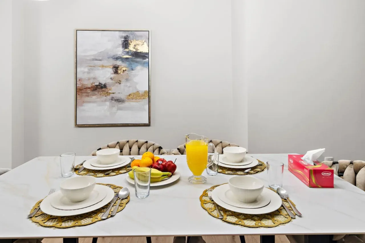 Una mesa de comedor está puesta con platos, individuales, fruta y un vaso de jugo de naranja.