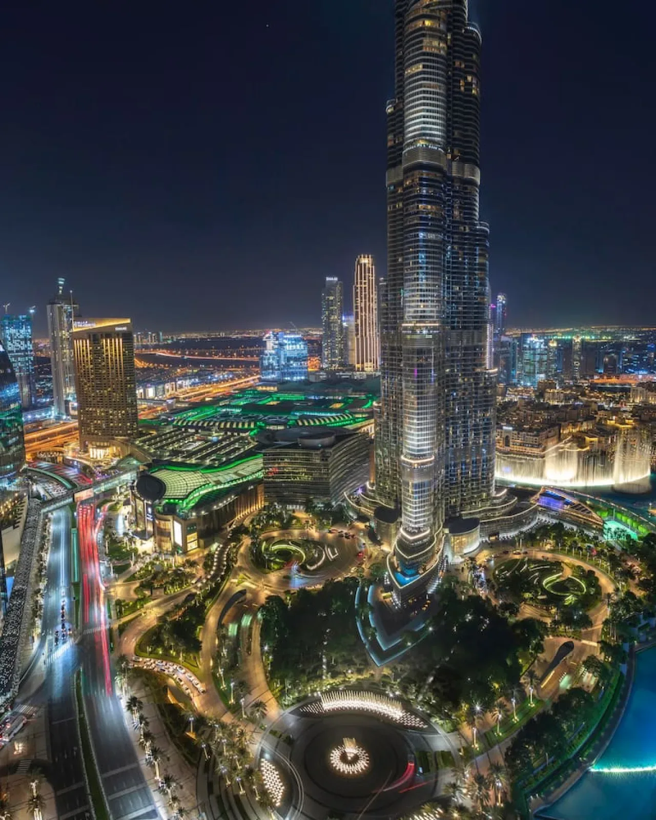 A nighttime city skyline with a tall illuminated skyscraper and landscaped grounds.