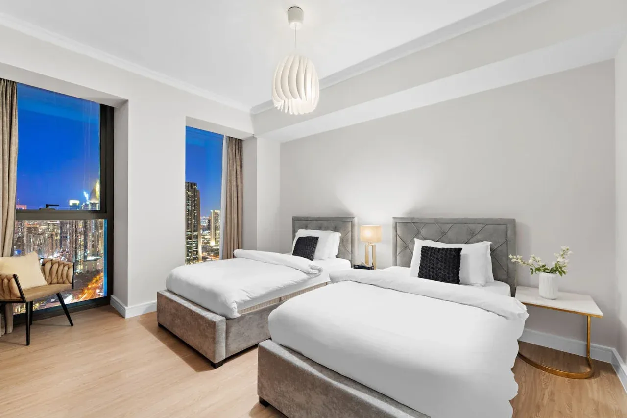 Un dormitorio luminoso con dos camas y grandes ventanas con vistas a las luces de la ciudad.