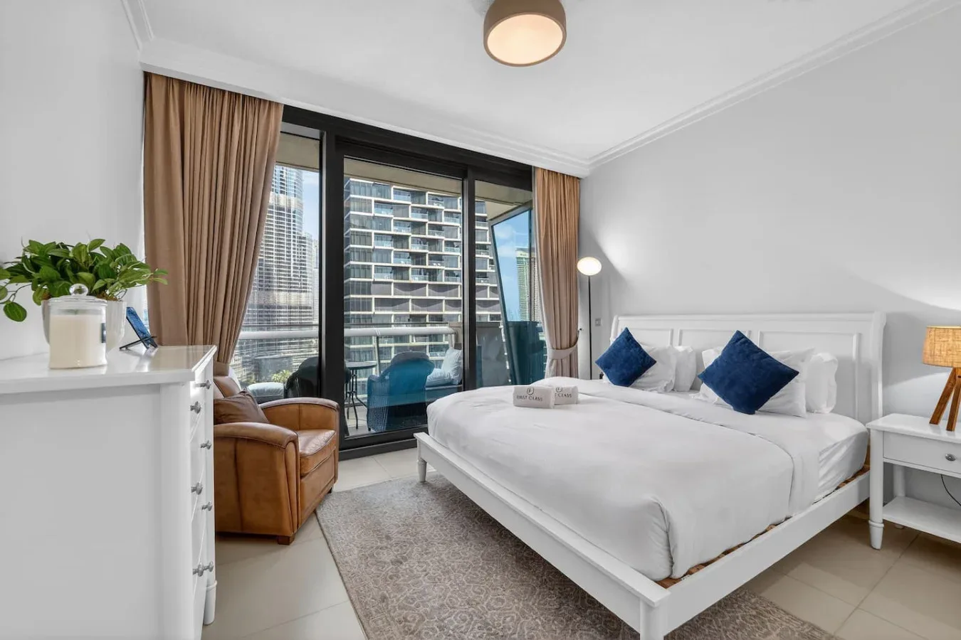 A bright bedroom with a white bed and large windows overlooking nearby buildings.