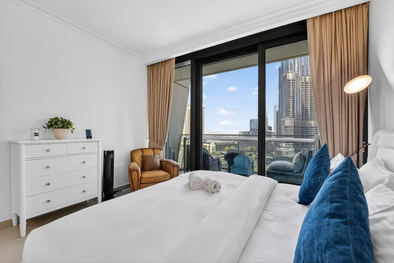 A bright bedroom with a white bed, blue cushions, and floor-to-ceiling windows.