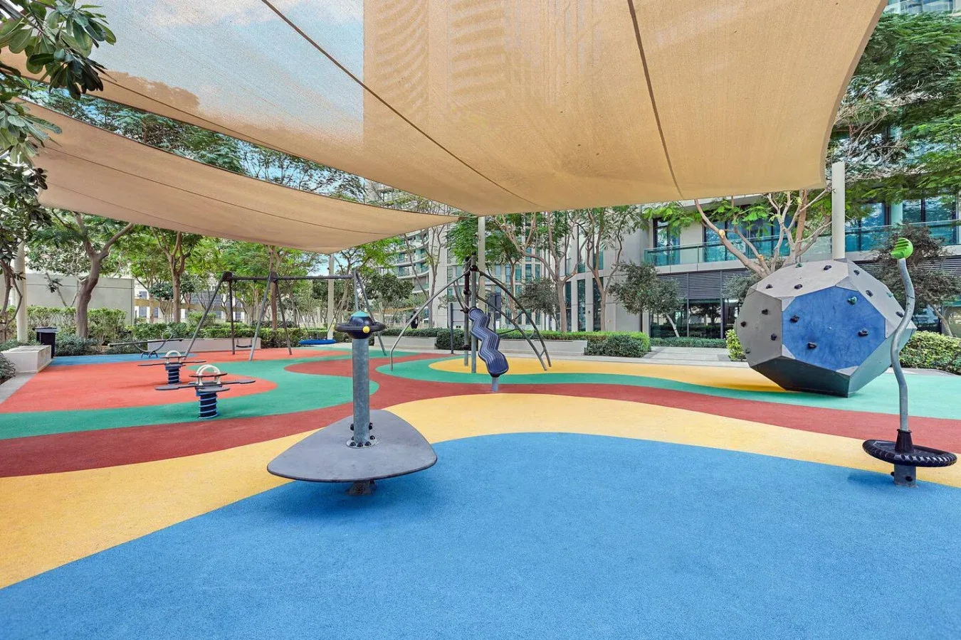 A colorful outdoor playground has soft play surfaces, climbing equipment, and shade canopies.
