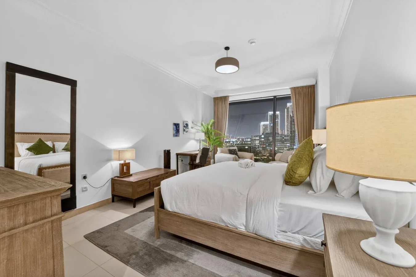 A bright bedroom with a large bed, bedside tables, and a window overlooking city lights.