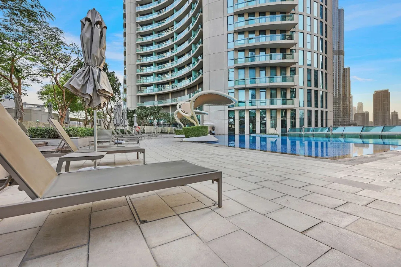 An outdoor pool deck with lounge seating beside tall modern buildings.