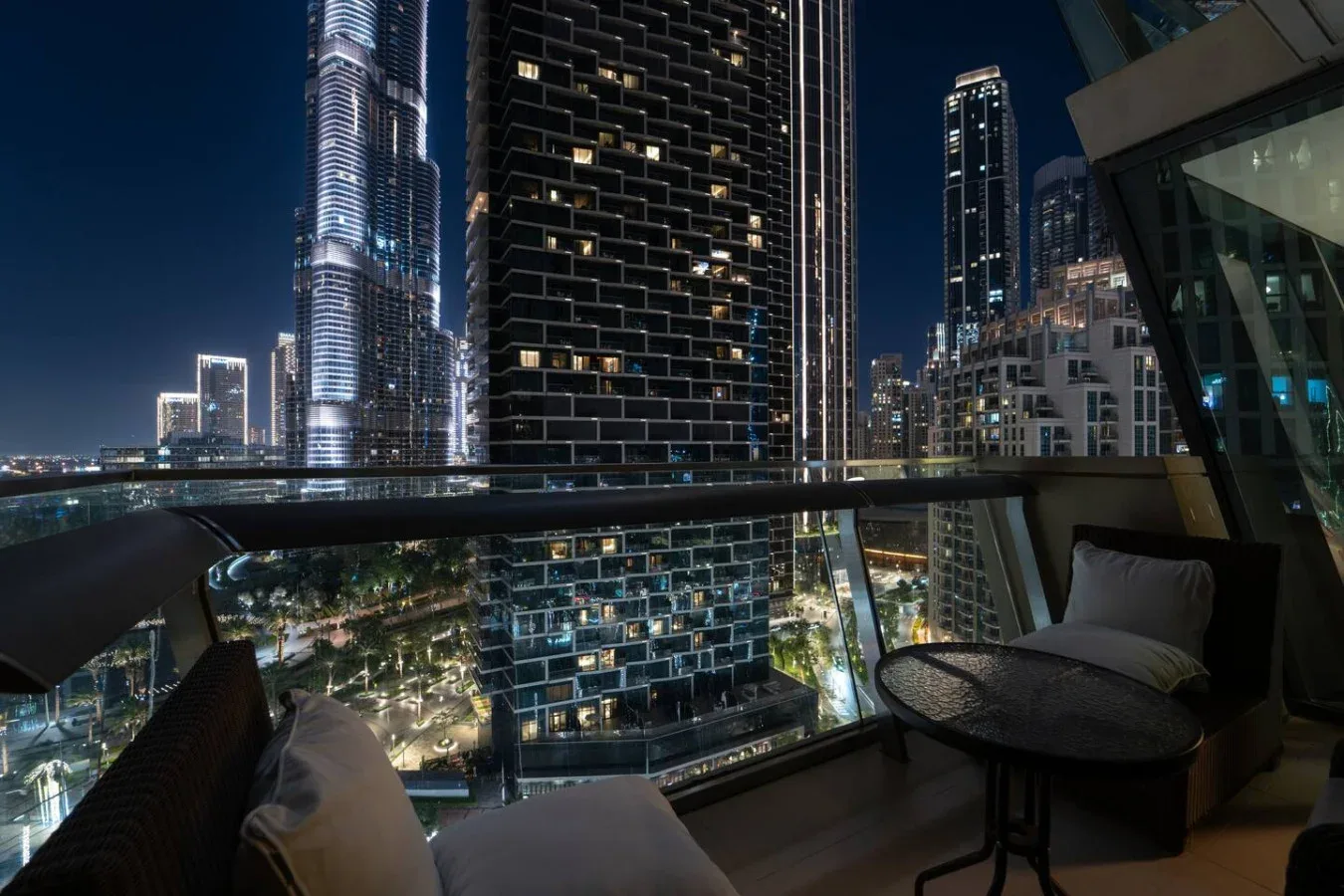 A nighttime balcony overlooks illuminated high-rise buildings, with seating and a small table.