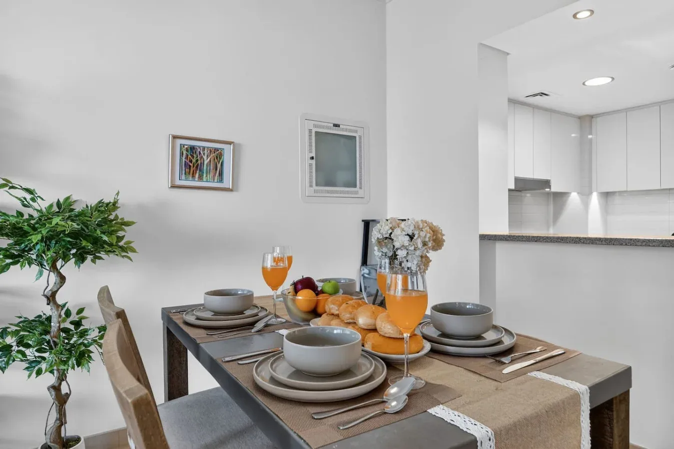 A dining table is set with plates and glasses beside a bright kitchen.