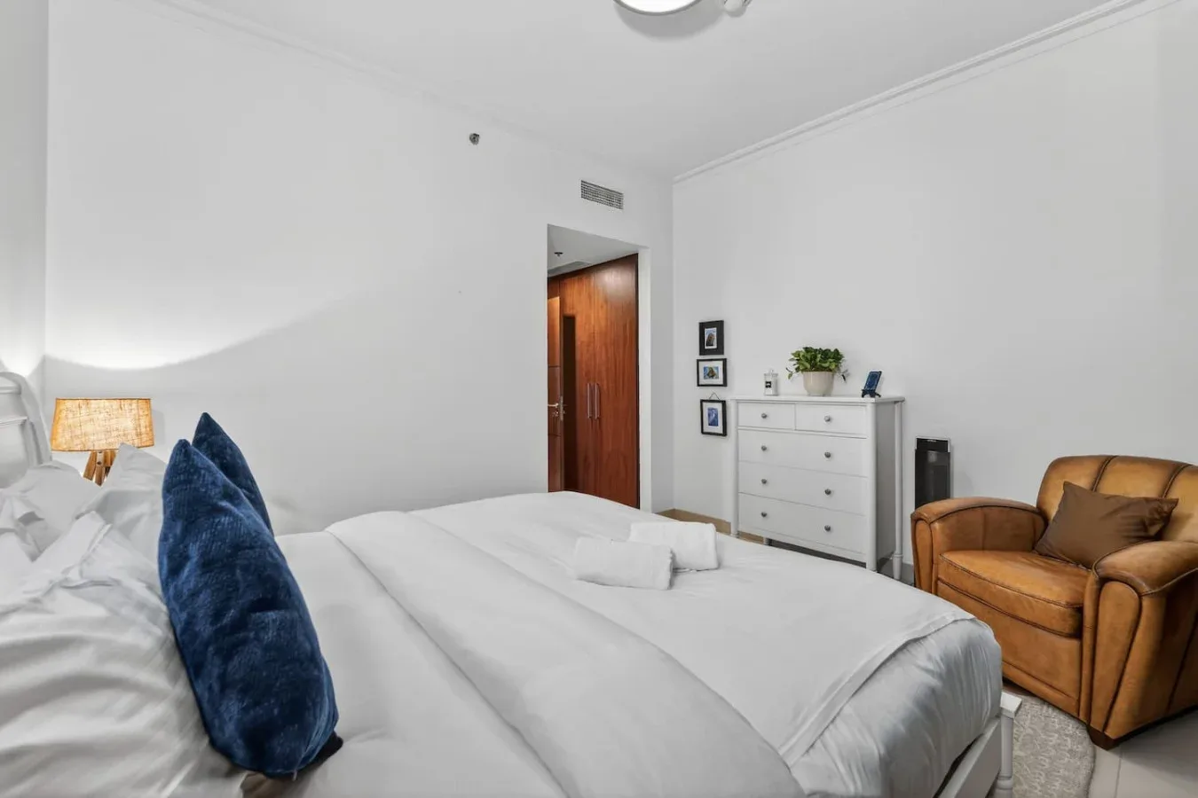 A bedroom with white bedding, a dresser, and an armchair.