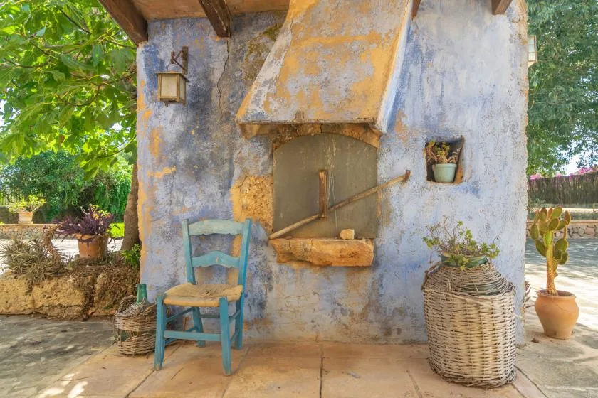 A rustic outdoor fireplace stands beside a blue chair and potted plants.