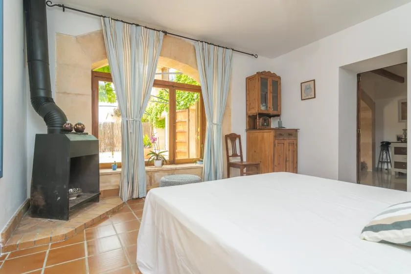 A bright bedroom with a large bed, curtained windows, and a black stove.
