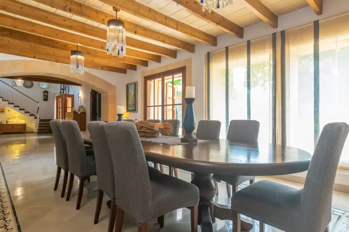 A dining area with a long table and upholstered seating beneath exposed wooden beams.