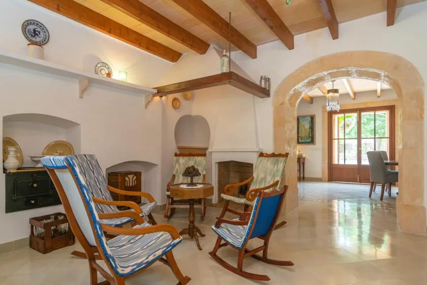 A bright living room with wooden seating and a dining area beyond the stone arch.