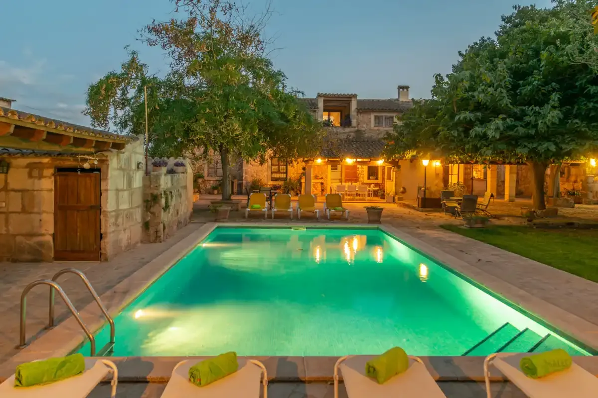 An illuminated swimming pool sits in a courtyard beside a stone building.