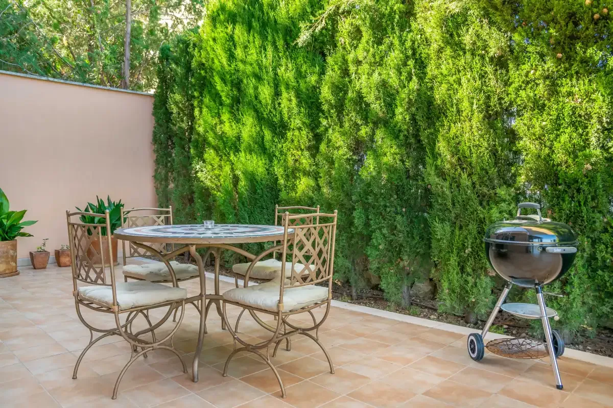 A tiled outdoor patio has a dining table and seating beside tall greenery.