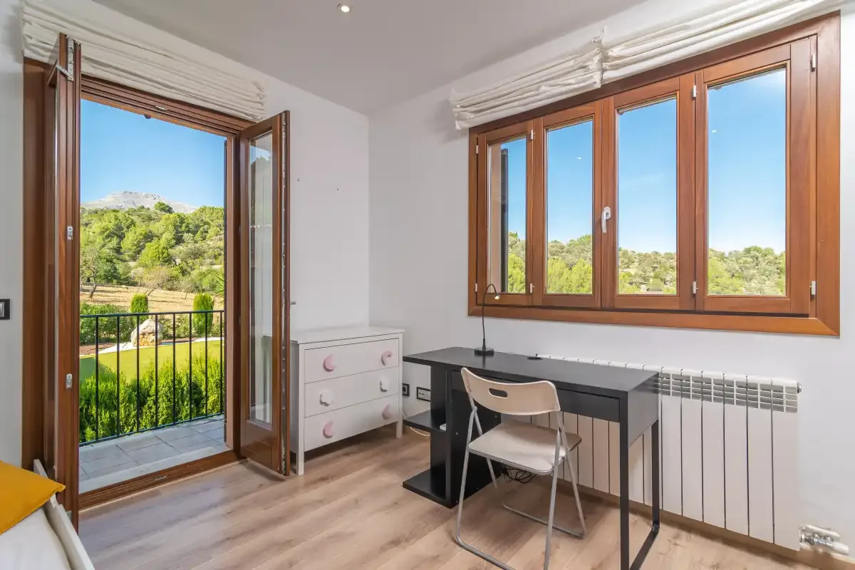 A bright room has a desk and chair beside open windows overlooking greenery.