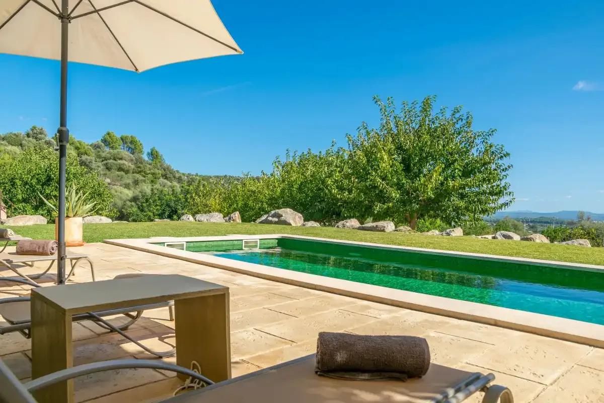 A swimming pool beside a paved patio with outdoor seating and an umbrella.