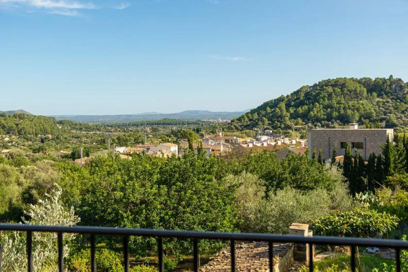 A balcony overlooks green hills, trees, and a cluster of buildings under a clear sky.
