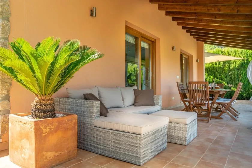 A covered patio has cushioned seating and a dining table beside the house.