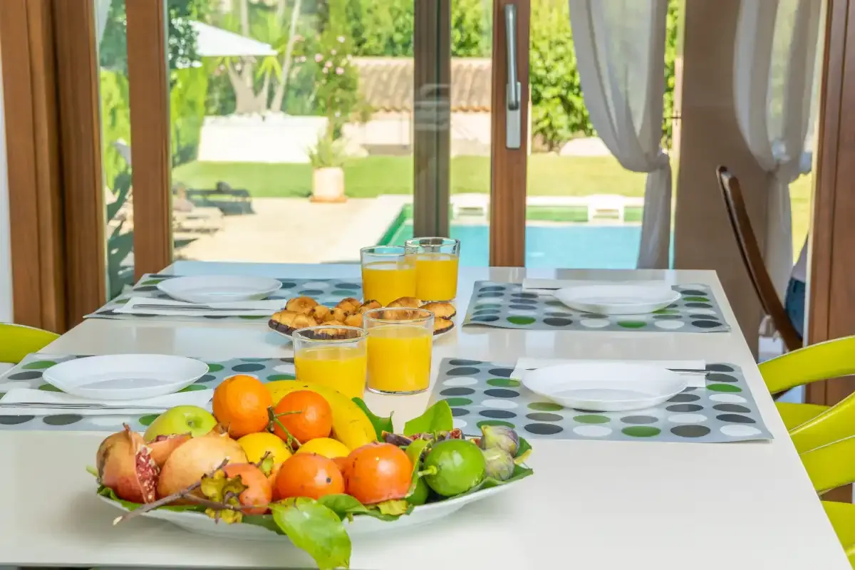 A dining table is set with fruit, pastries, and orange juice beside a garden view.