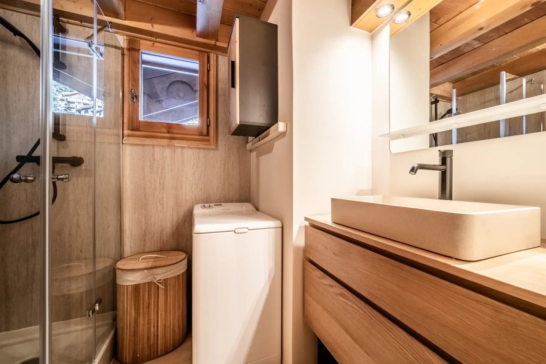 Modern bathroom with wooden accents, featuring a glass-enclosed shower, washing machine, and a vanity with a rectangular sink. A small window provides natural light.