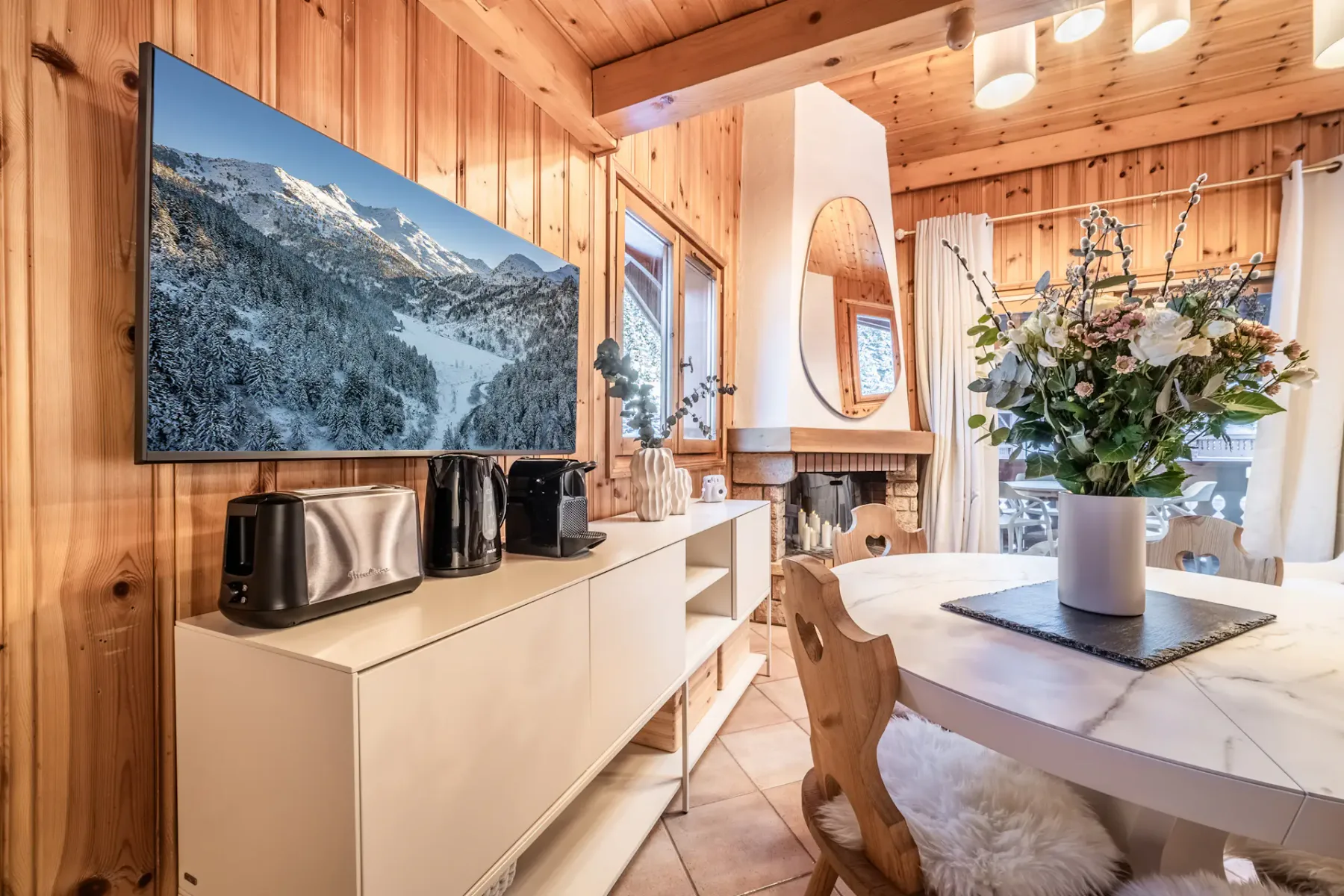 Wood-paneled kitchen with white cabinets, a small white shelf holding kitchen appliances, and a dining table with wooden chairs. A large window shows a snowy landscape outside.