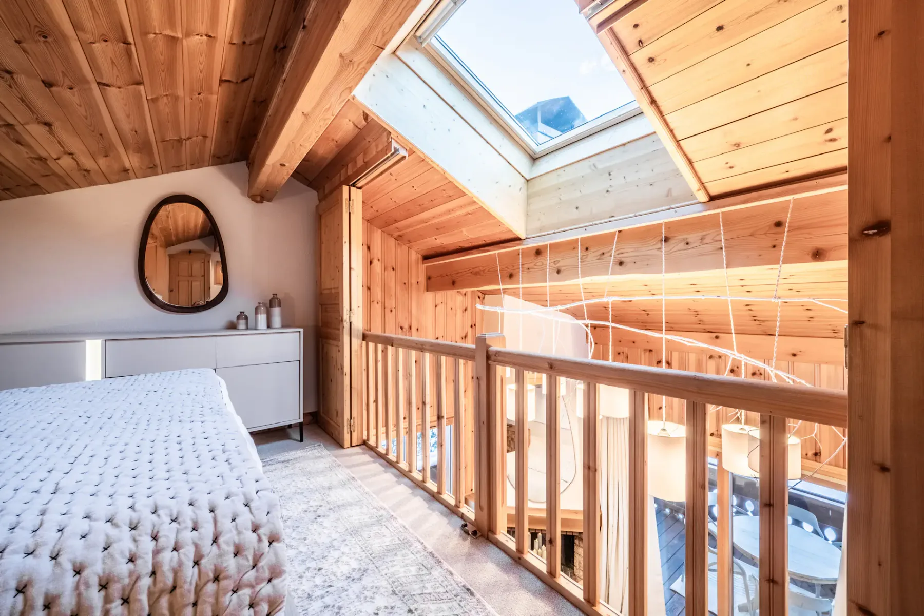 Cozy attic bedroom with wooden ceiling, double bed, round mirror on a white dresser, and a bedside lamp.