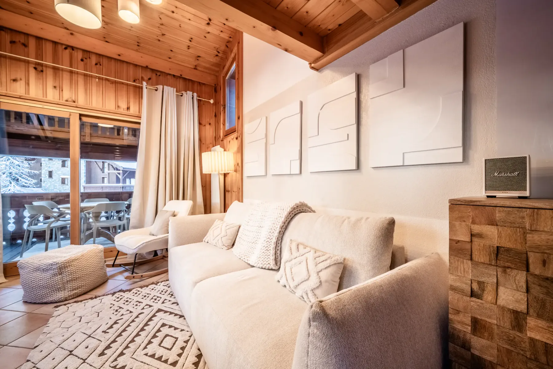 Cozy wooden living room with a round dining table, chairs, sofa, large windows, fireplace, textured rug, and a TV displaying a snowy mountain landscape.