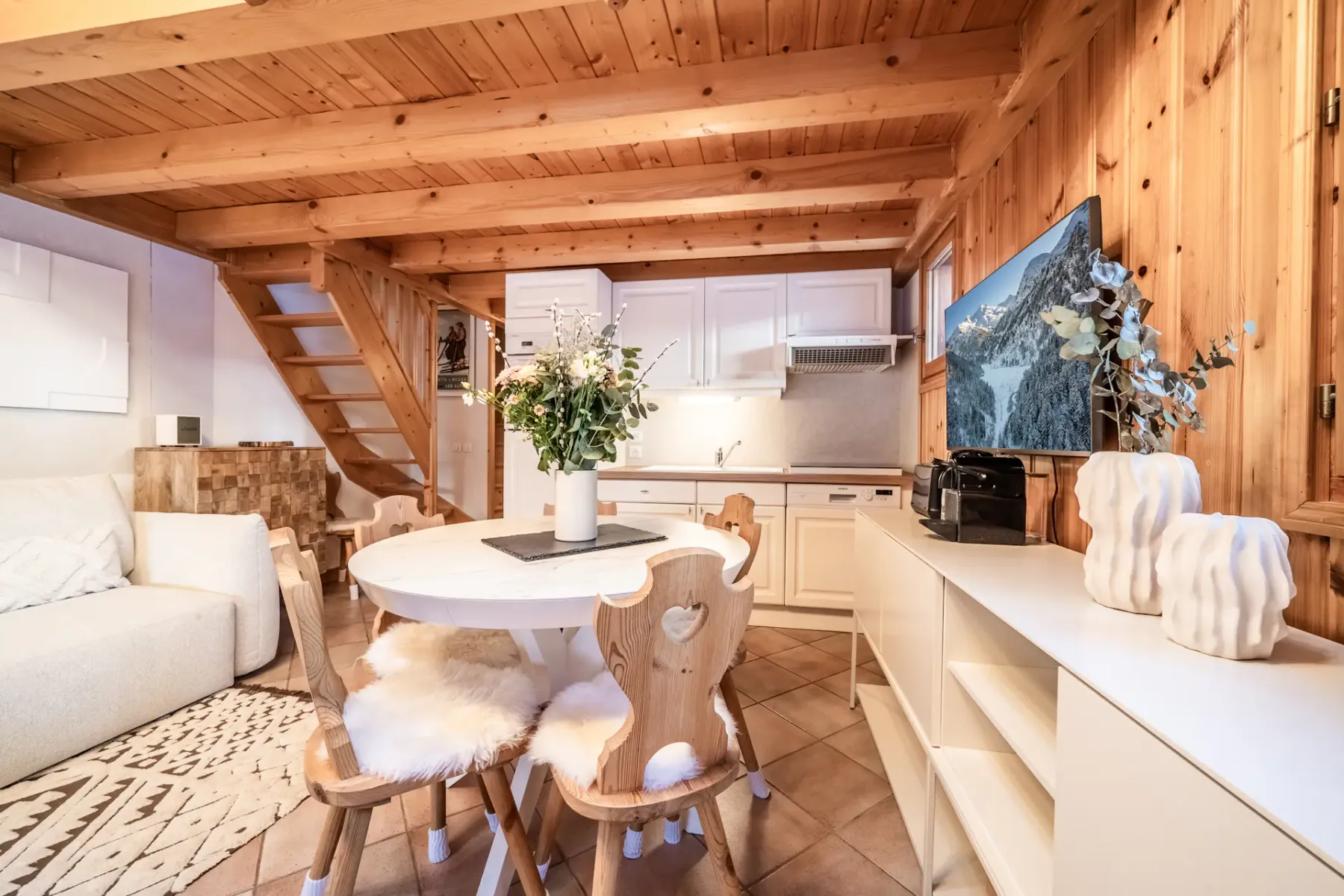 Cozy wooden chalet interior with a round dining table, fur-covered chairs, white cabinets, and a visible staircase. A vase with flowers and decorative items adorn the space.