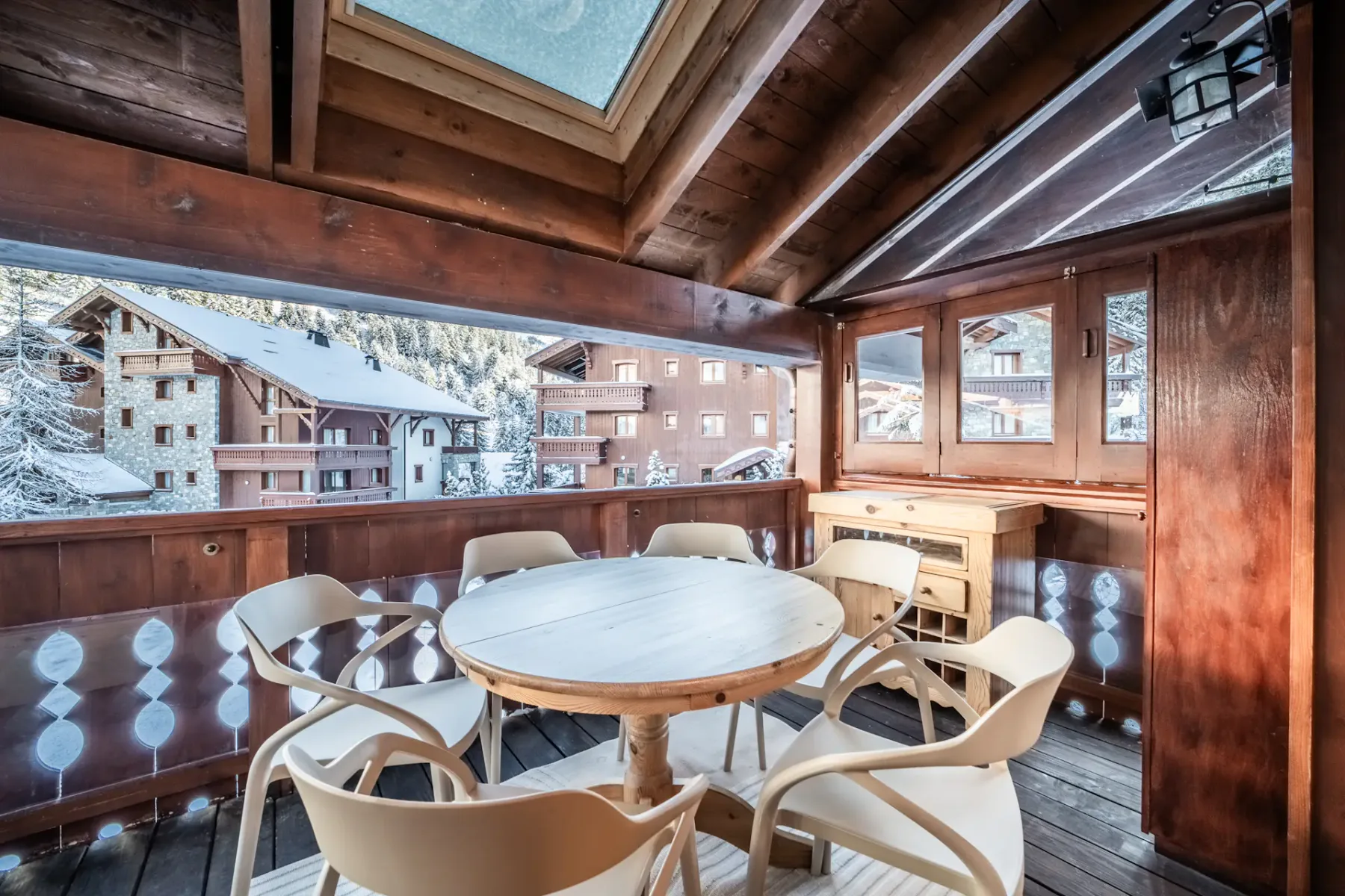 A cozy wooden balcony with a round table and six chairs overlooks a snowy alpine village with wooden buildings.