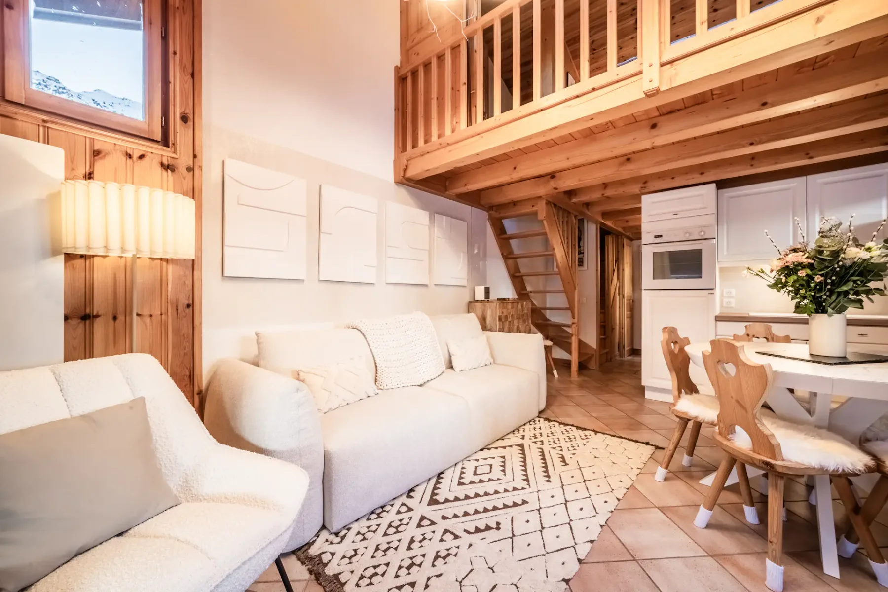 Cozy wooden living room with a round dining table, chairs, sofa, large windows, fireplace, textured rug, and a TV displaying a snowy mountain landscape.