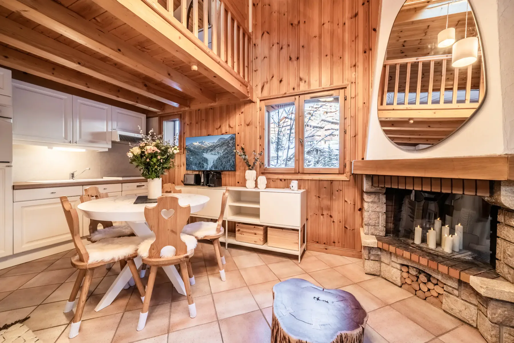 Cozy wooden chalet interior with a round dining table, fur-covered chairs, white cabinets, and a visible staircase. A vase with flowers and decorative items adorn the space.