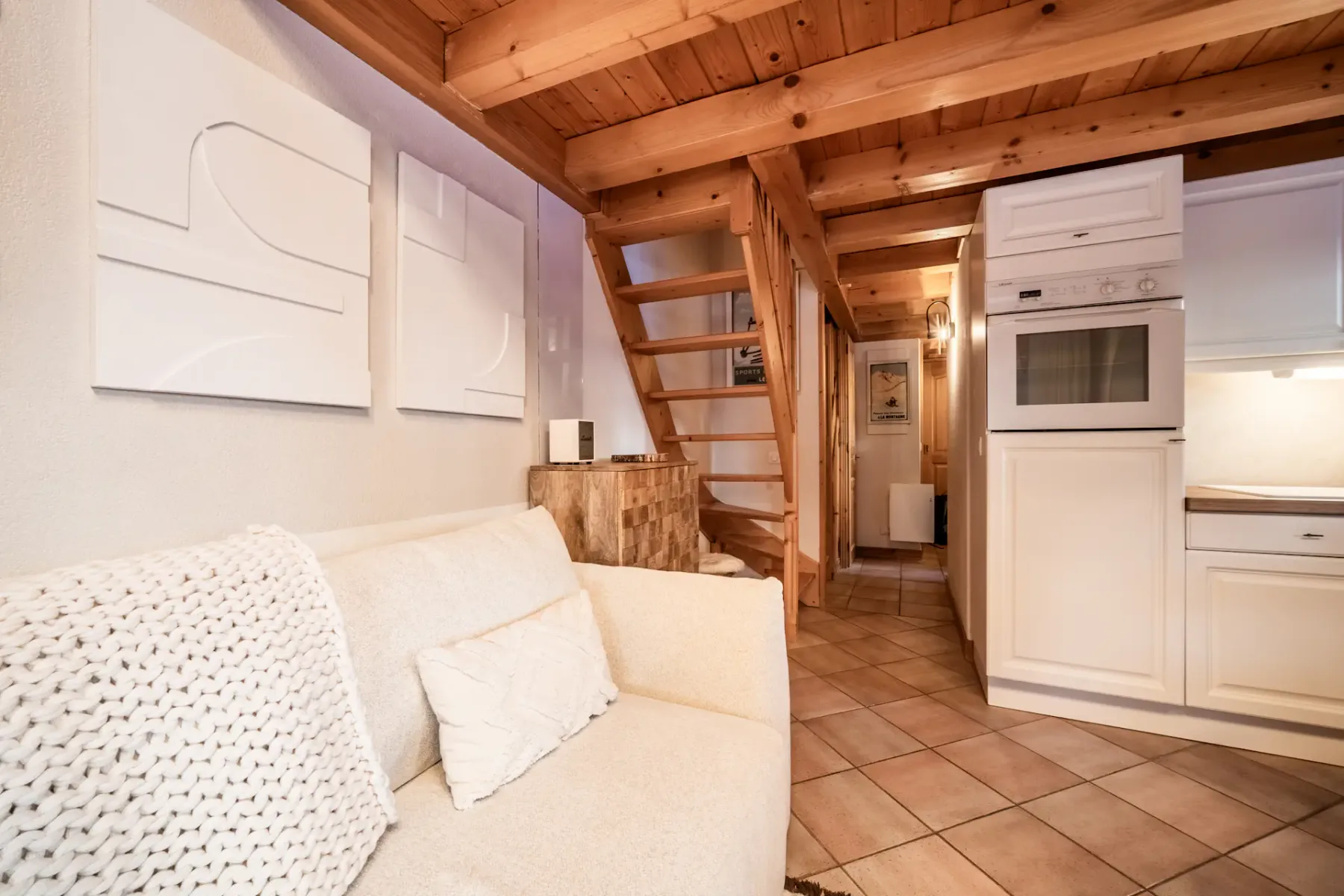 Small, cozy living area with a white sofa, neutral decor, wooden staircase, and a compact kitchen featuring white cabinets and built-in appliances.
