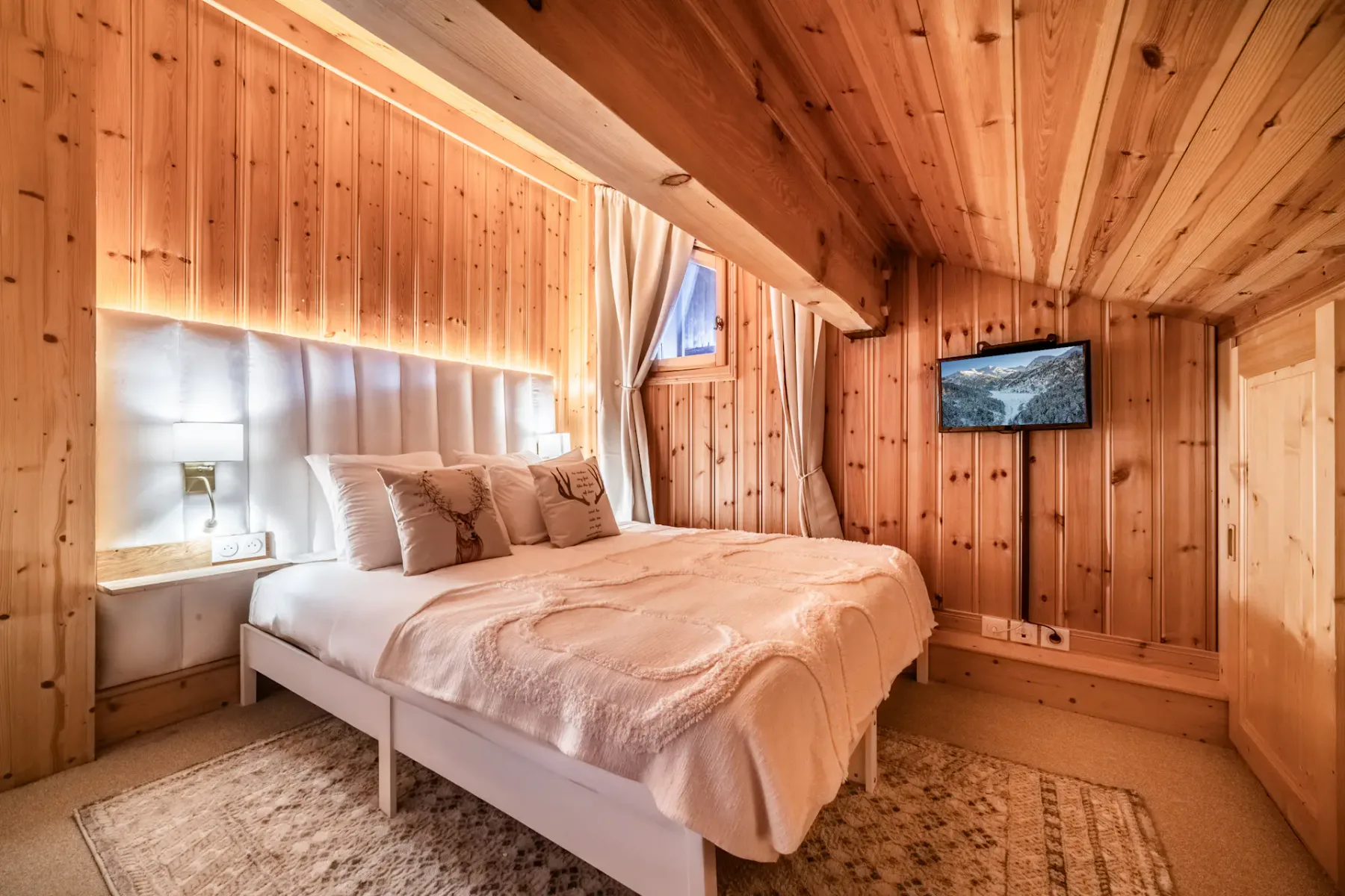 A cozy wooden cabin bedroom with a double bed, soft lighting, and a mounted TV. The room features a window with curtains and rustic decor.