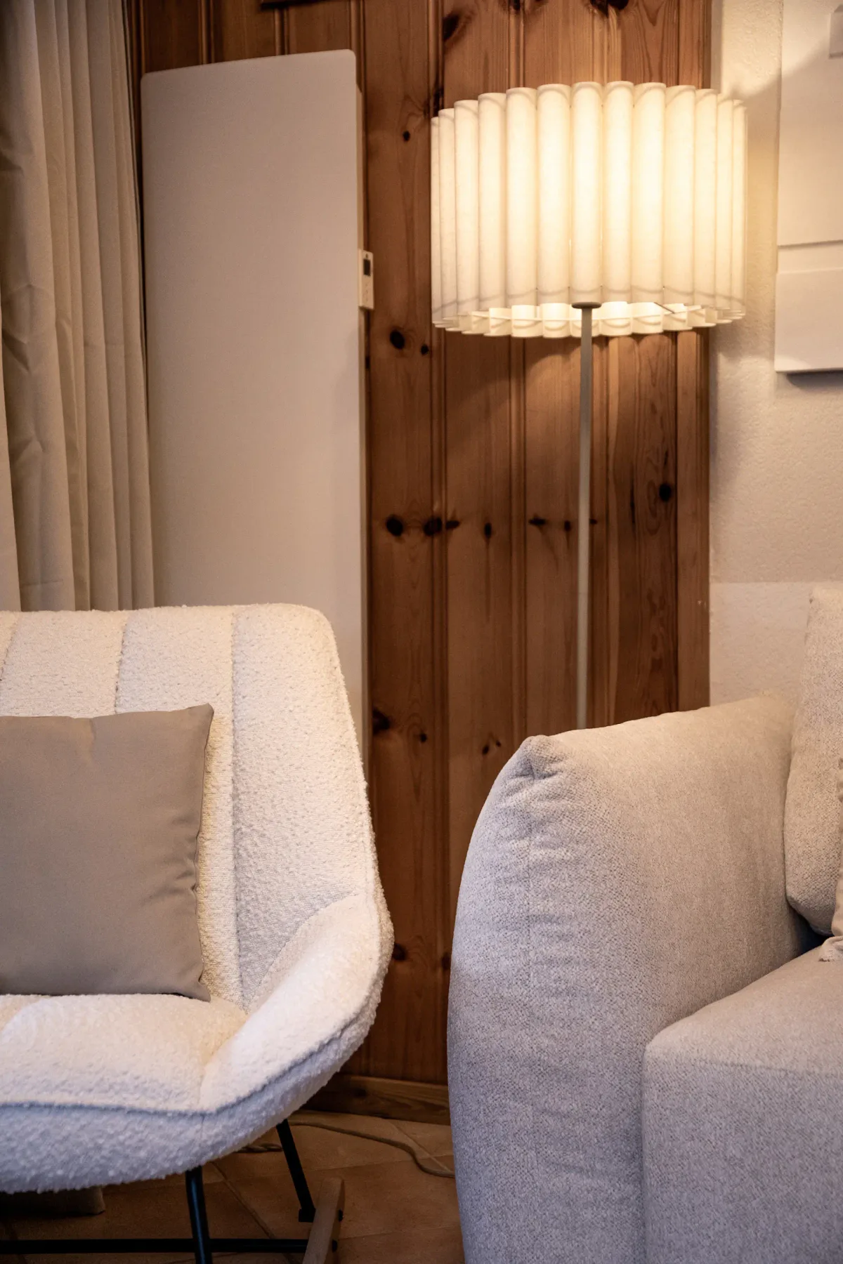 Cozy living room featuring a white cushioned chair, a soft beige couch, and a tall, cylindrical lampshade beside a wooden wall.