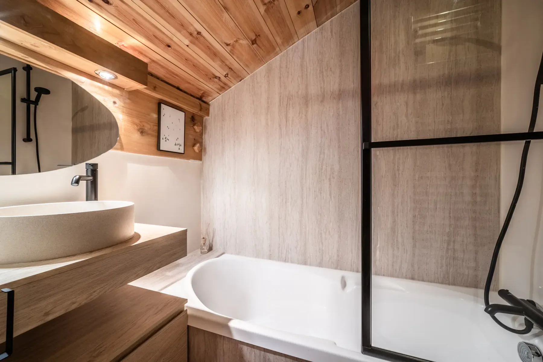 A modern bathroom with a wooden ceiling, wall mirror, round sink, and a bathtub with a glass partition.