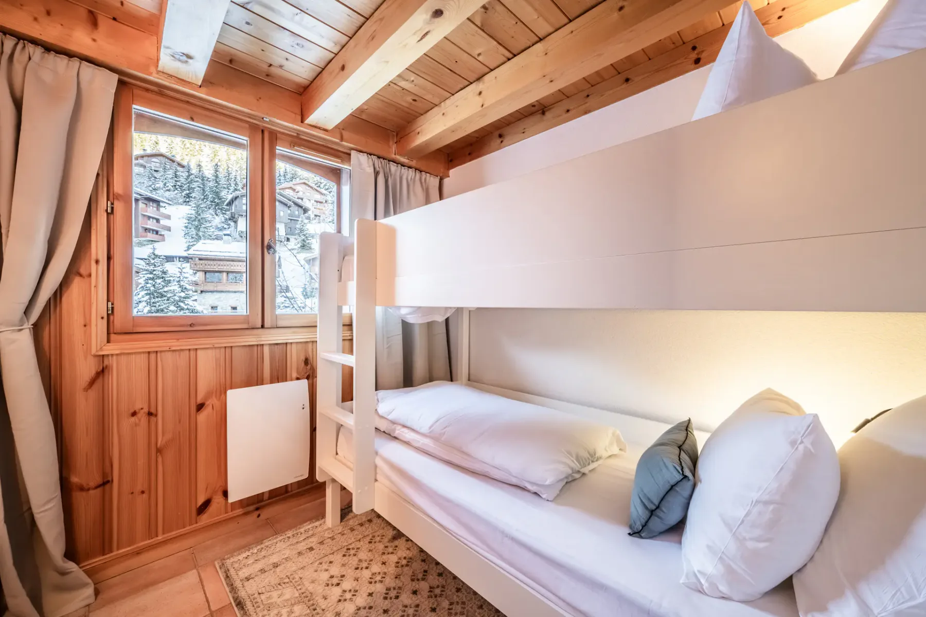 Cozy room with bunk beds, a heart-decorated door, wooden beams, and a window with a view of snow-covered houses.