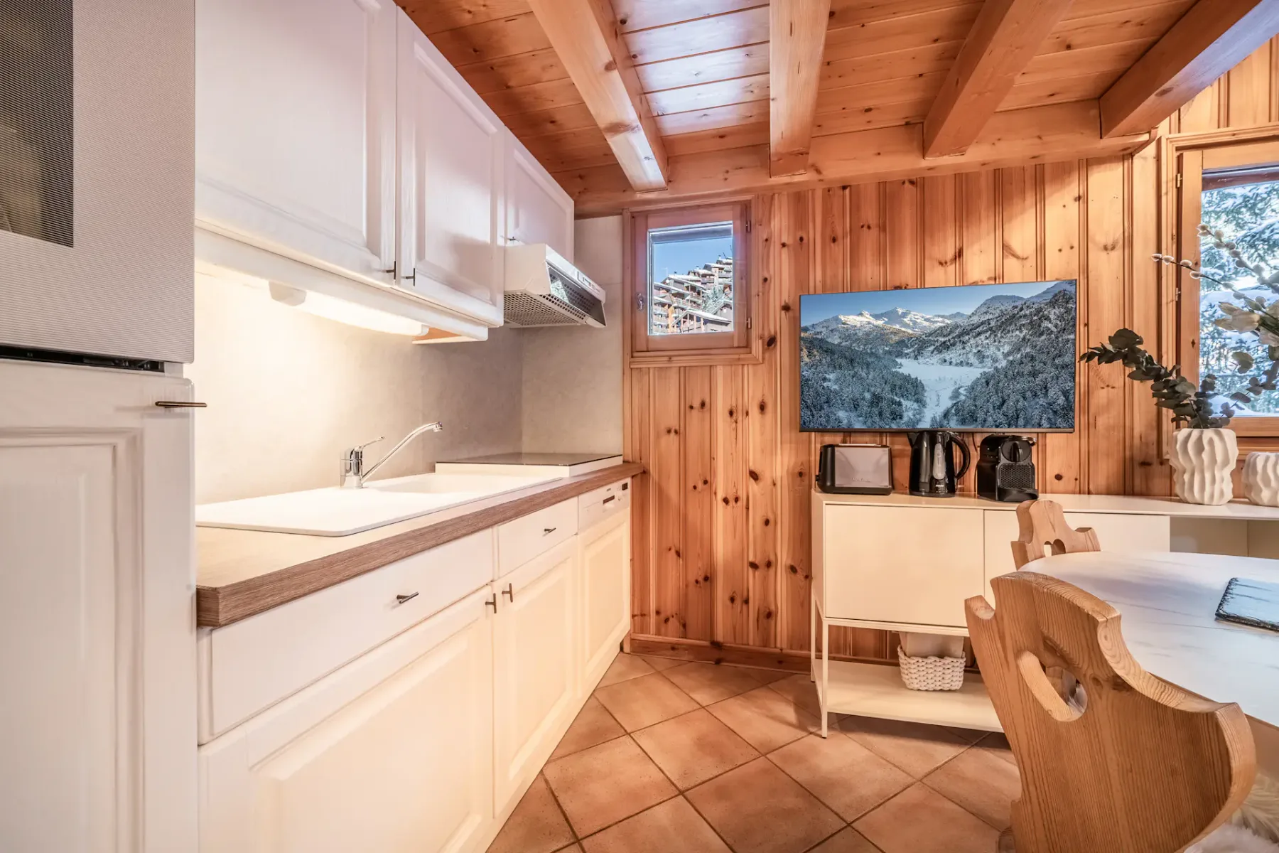 Wood-paneled kitchen with white cabinets, a small white shelf holding kitchen appliances, and a dining table with wooden chairs. A large window shows a snowy landscape outside.