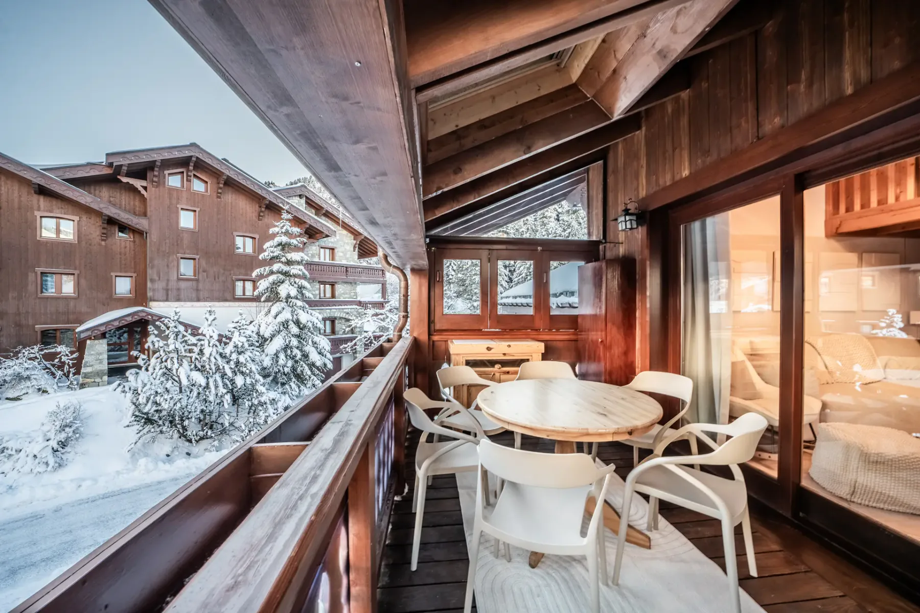 A cozy wooden balcony with a round table and six chairs overlooks a snowy alpine village with wooden buildings.
