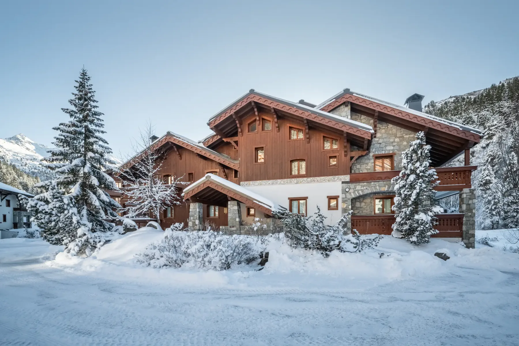A large wooden chalet surrounded by snow-covered trees and shrubs on a clear winter day.
