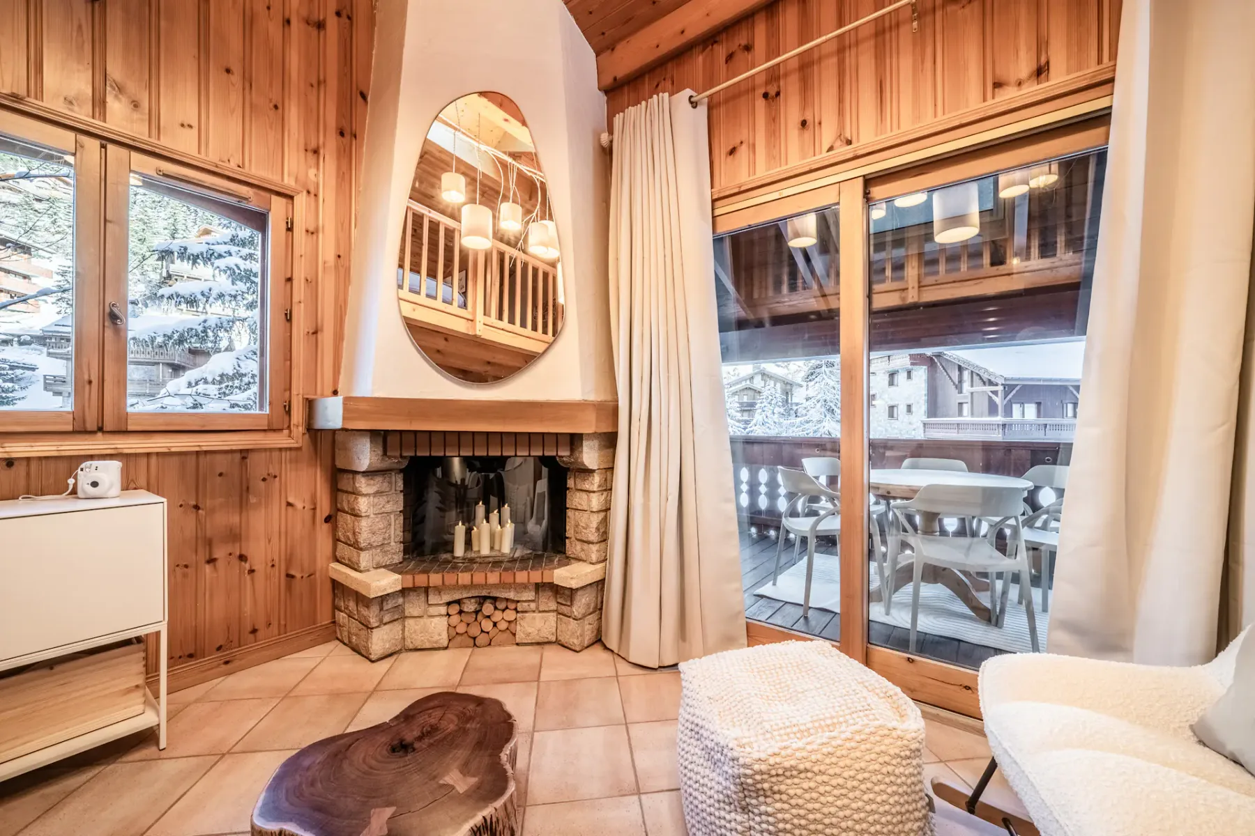 Cozy wooden living room with a round dining table, chairs, sofa, large windows, fireplace, textured rug, and a TV displaying a snowy mountain landscape.