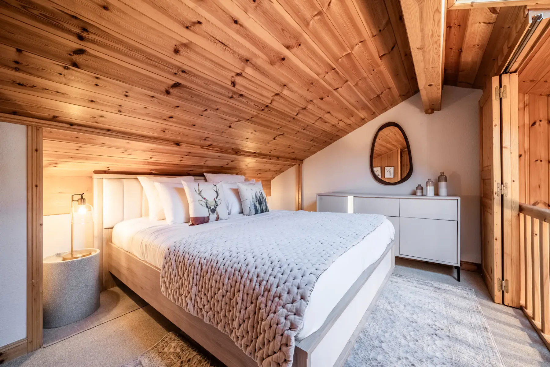 Cozy attic bedroom with wooden ceiling, double bed, round mirror on a white dresser, and a bedside lamp.