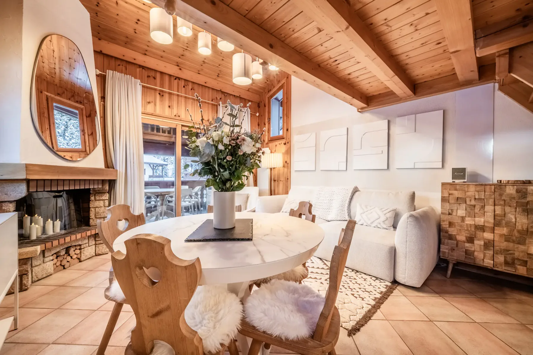 Cozy wooden chalet interior with a round dining table, fur-covered chairs, white cabinets, and a visible staircase. A vase with flowers and decorative items adorn the space.