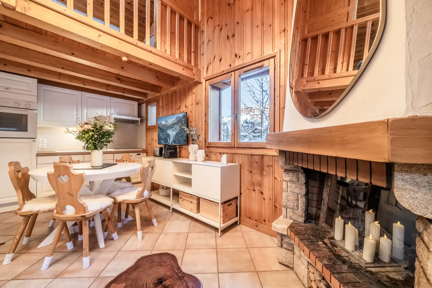 Cozy wooden chalet interior with a round dining table, fur-covered chairs, white cabinets, and a visible staircase. A vase with flowers and decorative items adorn the space.