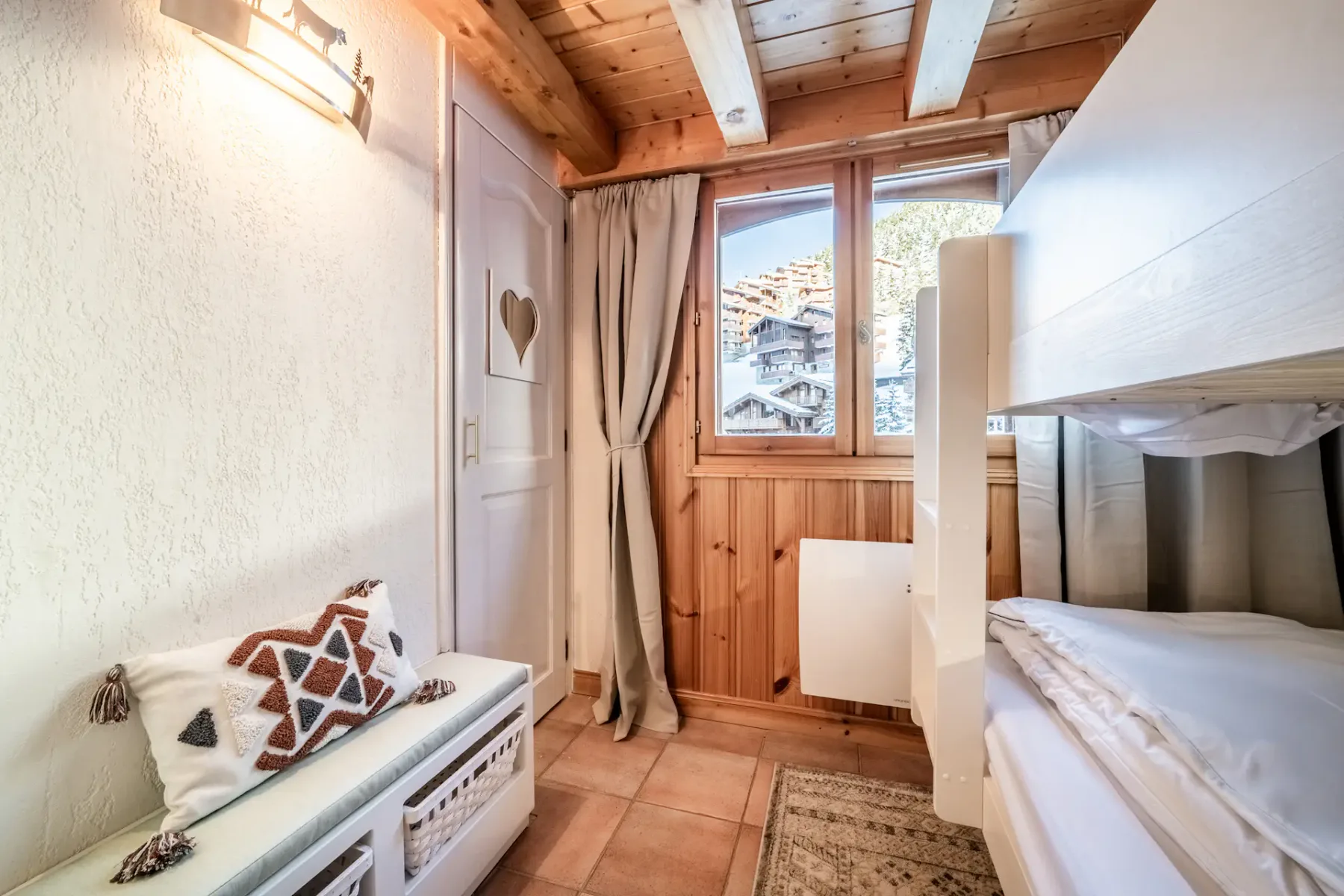Cozy room with bunk beds, a heart-decorated door, wooden beams, and a window with a view of snow-covered houses.