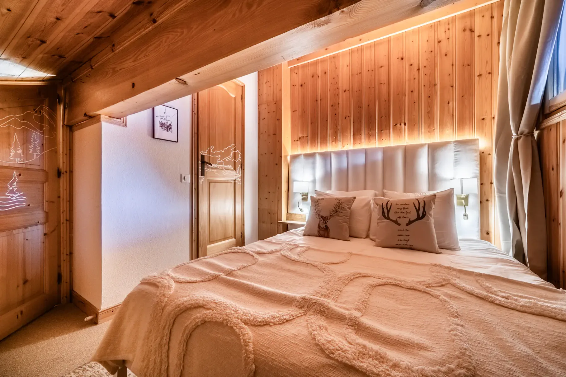 A cozy wooden cabin bedroom with a double bed, soft lighting, and a mounted TV. The room features a window with curtains and rustic decor.