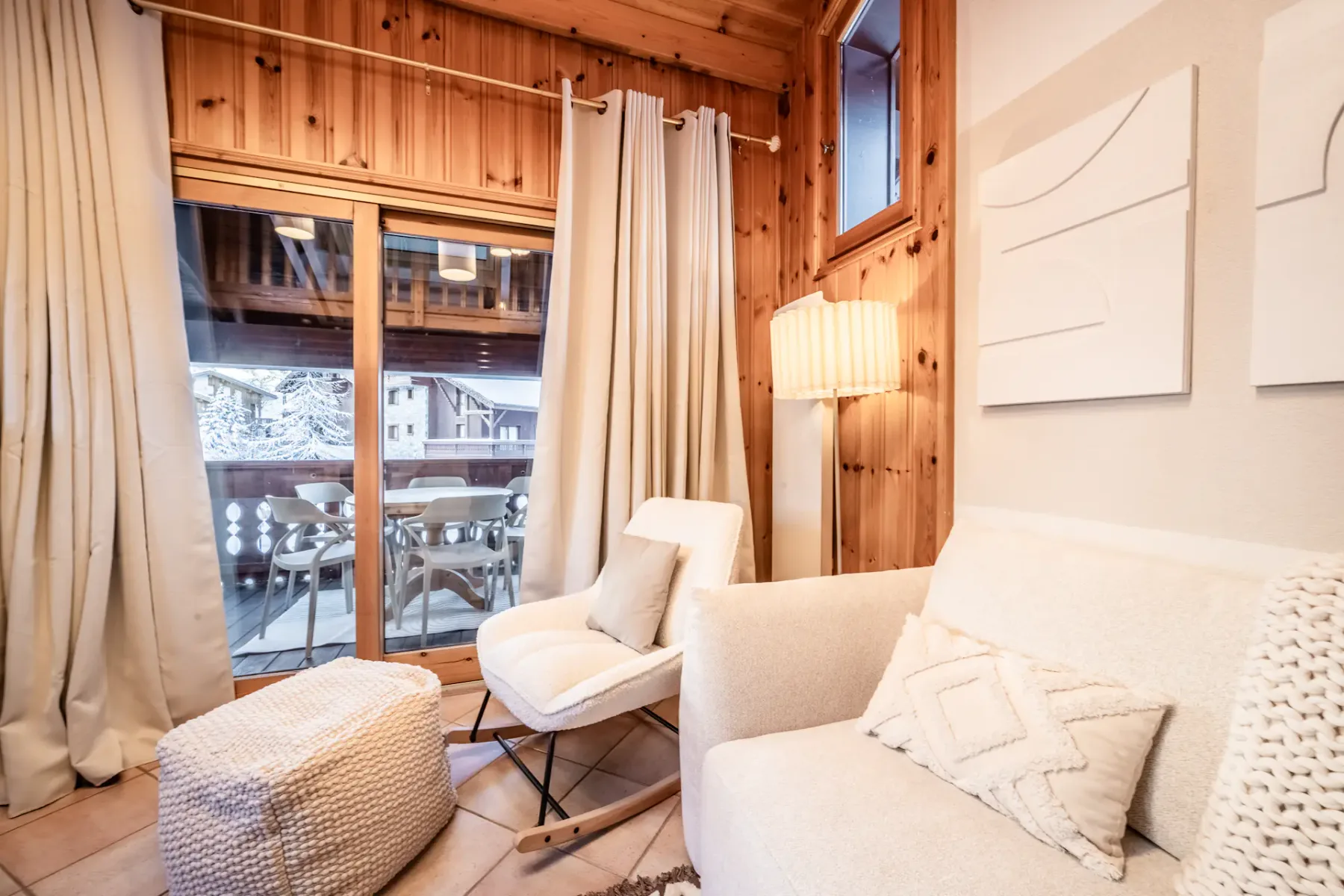 Cozy wooden living room with a round dining table, chairs, sofa, large windows, fireplace, textured rug, and a TV displaying a snowy mountain landscape.