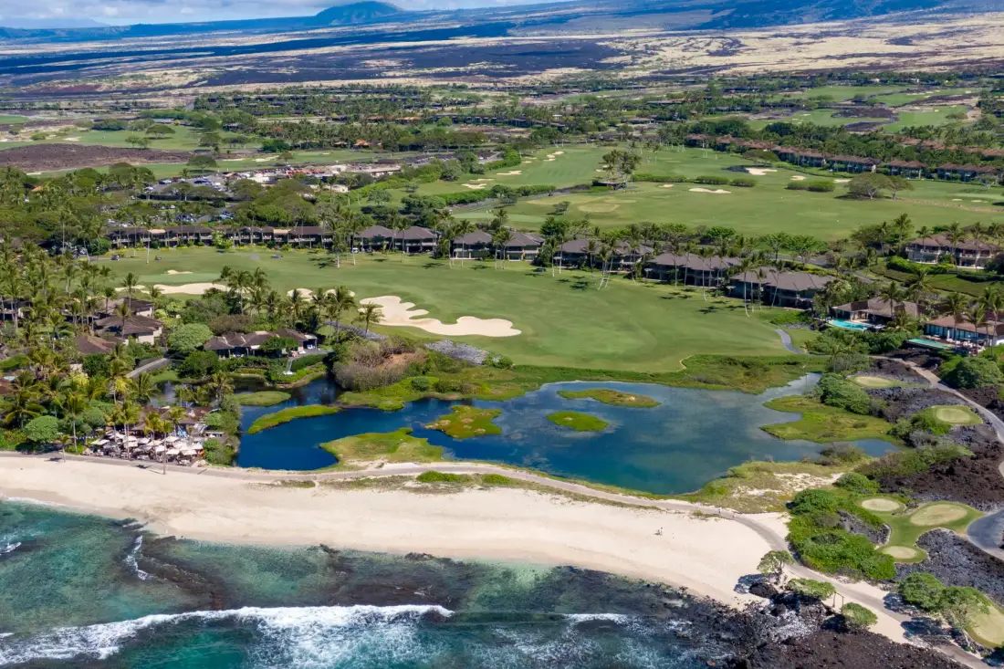 An aerial view shows a coastal golf course, ponds, and a sandy beach.