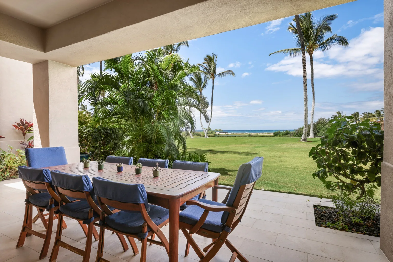 A covered patio has a dining table and chairs overlooking a lawn and ocean.